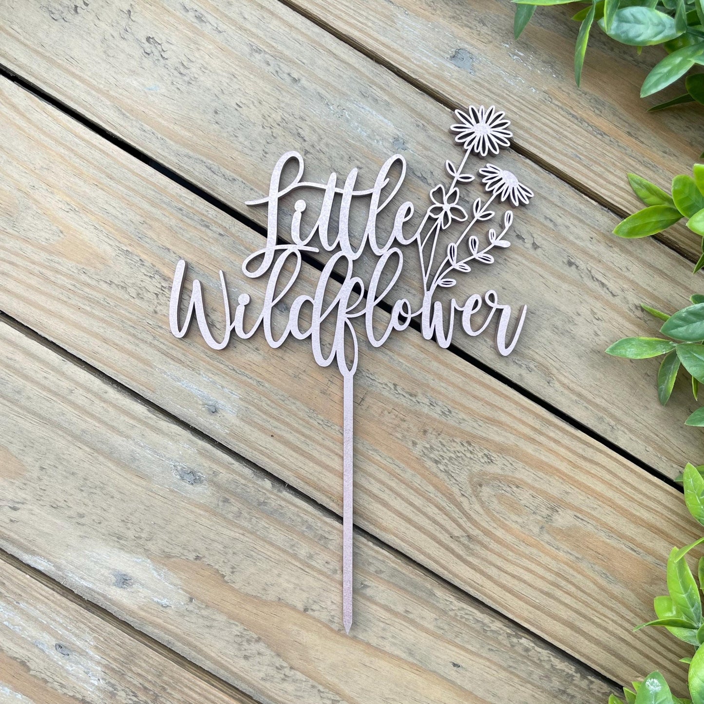 May include: A wooden cake topper with the words "little wildflower" and a floral design. The topper is light brown and has a stick for inserting into a cake.