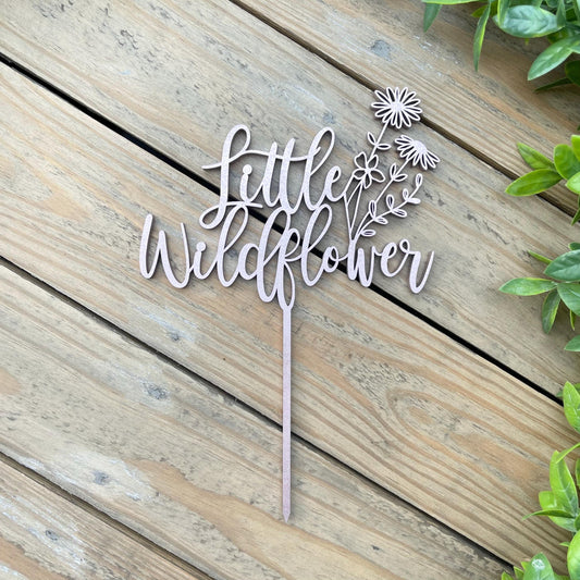 May include: A wooden cake topper with the words "little wildflower" and a floral design. The topper is light brown and has a stick for inserting into a cake.